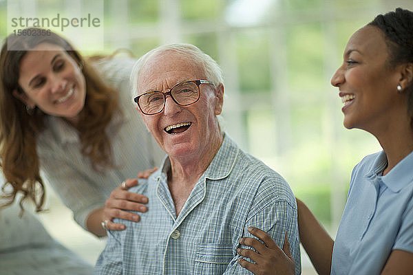 Porträt eines lachenden älteren Mannes  der von einer Krankenschwester und seiner erwachsenen Tochter getröstet wird  während er einen Pyjama trägt.