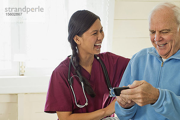 Ein älterer Mann benutzt ein Mobiltelefon  während eine lächelnde junge Krankenschwester zuschaut