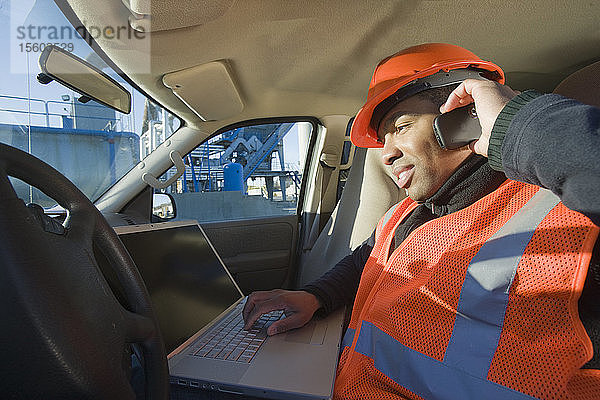 Ingenieur  der in einem Auto an einem Laptop arbeitet und mit einem Mobiltelefon telefoniert