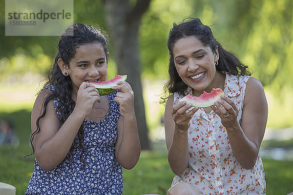 Glückliche hispanische Mutter mit ihrer Teenager-Tochter  die im Park eine Wassermelone isst