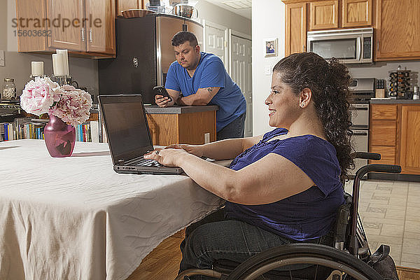 Frau mit Spina Bifida am Computer mit ihrem Mann beim Telefonieren zu Hause