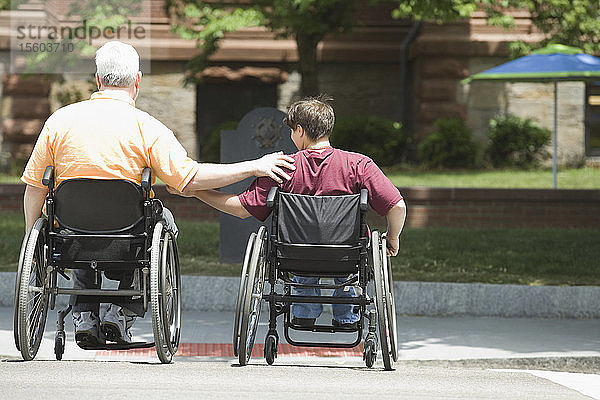 Rückansicht eines Mannes mittleren Alters und einer Frau mittleren Alters mit Muskeldystrophie  die in Rollstühlen eine Straße überqueren
