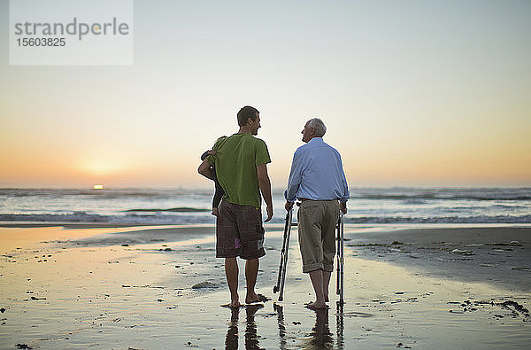 Älterer Mann mit einer Gehhilfe am Strand mit seinem Sohn und seinem Enkelkind