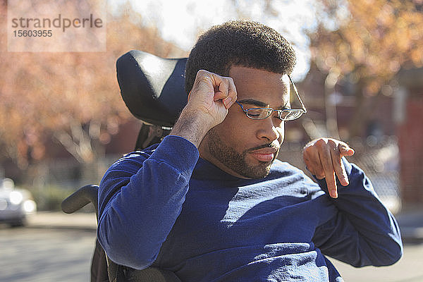 Afroamerikanischer Mann mit Cerebralparese  der draußen auf seinem Elektrorollstuhl eine Brille aufsetzt