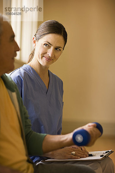 Eine Krankenschwester lächelt  während sie mit einem Mann zusammensitzt  der ein Handgewicht benutzt