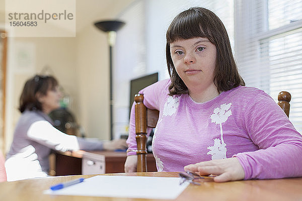 Mädchen mit Down-Syndrom sitzt auf einem Stuhl  während ihre Mutter im Hintergrund einen Computer benutzt