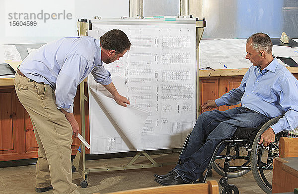Zwei Projektingenieure benutzen ein Hängeregal für Baustellenpläne  einer sitzt im Rollstuhl und hat eine Wirbelsäulenverletzung