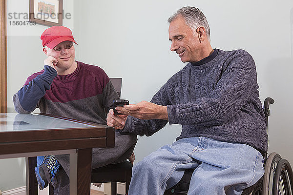 YJunger Mann mit Down-Syndrom und sein Vater im Rollstuhl mit Rückenmarksverletzung benutzen zu Hause ein Mobiltelefon