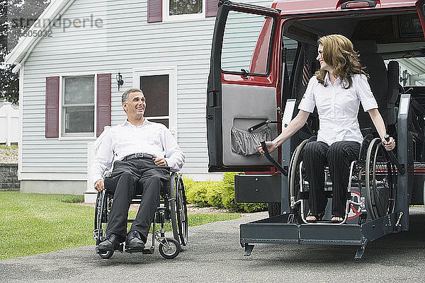 Älterer Mann  der mit einer Rückenmarksverletzung in einem Rollstuhl sitzt und auf eine mittelgroße erwachsene Frau mit einer Rückenmarksverletzung blickt  die von einem Rollstuhllift heruntergelassen wird