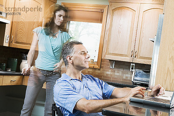 Seitenprofil eines mittelgroßen Mannes  der im Rollstuhl sitzt und einen Laptop benutzt  während eine mittelgroße Frau hinter ihm geht