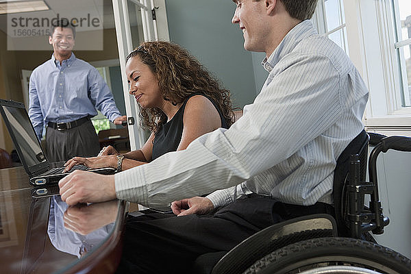 Ein Geschäftsmann mit einer Rückenmarksverletzung und eine sehbehinderte hispanische Geschäftsfrau benutzen einen Laptop in einem Büro