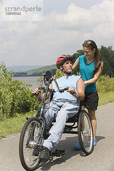 Junger Mann mit einer Rückenmarksverletzung fährt adaptives Fahrrad mit einer jungen Frau  die hinter ihm steht und lächelt