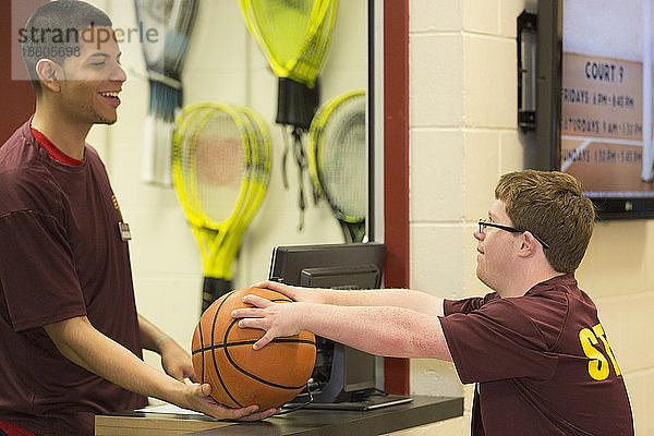 Junger Mann mit Down-Syndrom hält einen Basketball mit einem Betreuer in der Ausgabestelle für College-Ausrüstung