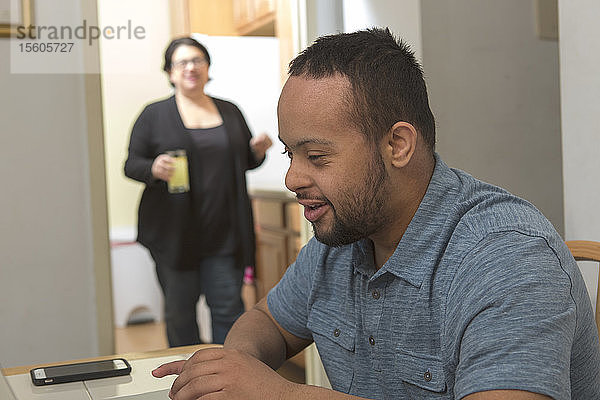 Afroamerikanischer Mann mit Down-Syndrom benutzt zu Hause einen Laptop  im Hintergrund seine Mutter