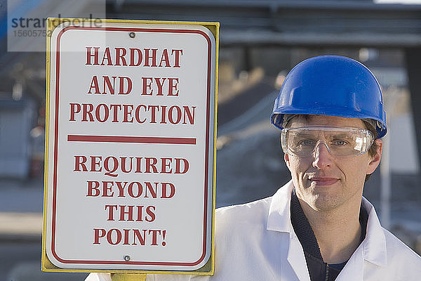 Laborarbeiter neben einem Warnschild mit der Aufschrift Hardhat and Eye Protection Required Beyond This Point in einem Asphaltwerk