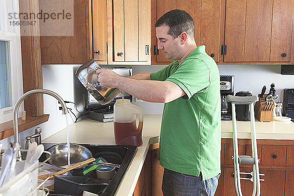 Mann nach Operation des vorderen Kreuzbandes (ACL) mit Krücken beim Teekochen und Abwaschen in der Küche