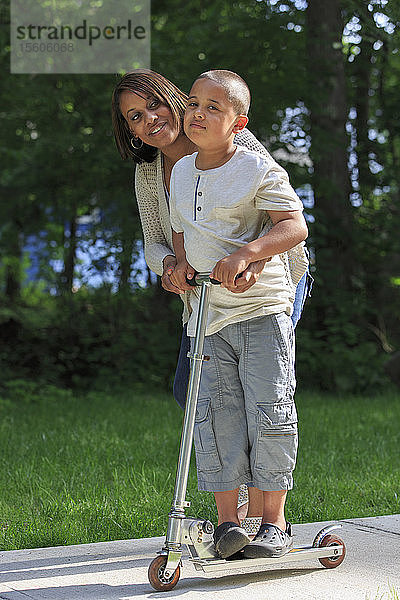 Hispanischer Junge mit Autismus  der mit seiner Mutter im Park auf einem Tretroller spielt