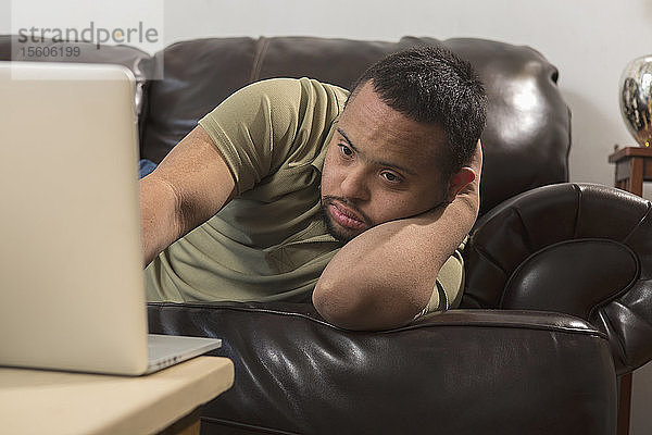 Afroamerikanischer Mann mit Down-Syndrom  der zu Hause vom Sofa aus einen Laptop benutzt