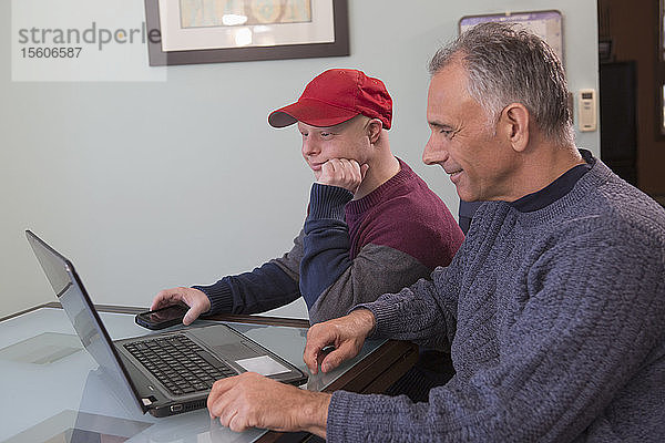 Junger Mann mit Down-Syndrom und sein Vater mit Rückenmarksverletzung benutzen zu Hause einen Laptop
