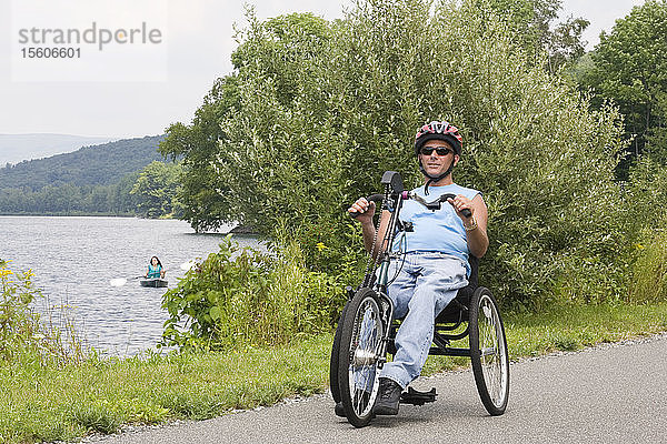 Junger Mann mit einer Rückenmarksverletzung auf einem adaptiven Fahrrad mit einer jungen Frau  die auf einem See Kajak fährt  im Hintergrund