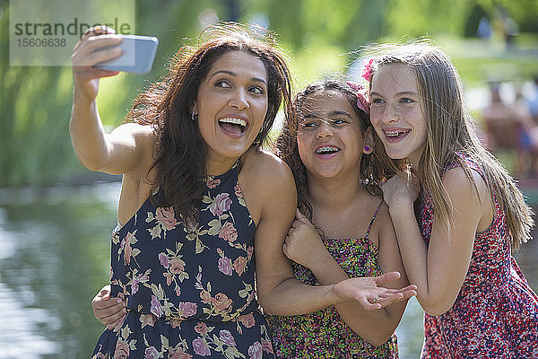 Glückliche hispanische Mutter mit zwei Teenager-Töchtern mit Zahnspange  die ein Selfie mit einem Mobiltelefon im Park machen