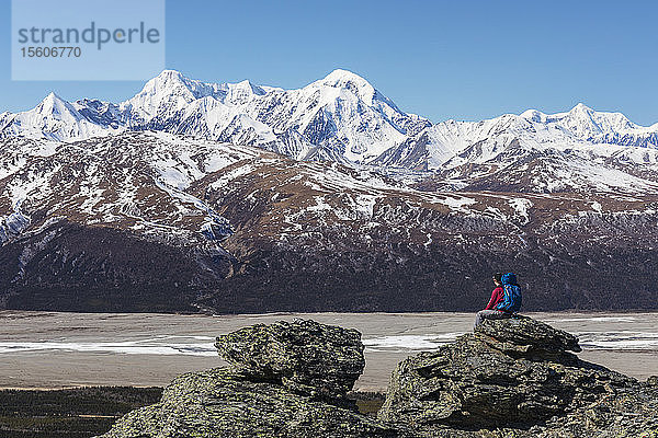 Ein Wanderer genießt die Aussicht auf McGinnis Peak  Mount Moffit und Mount Hayes in der Eastern Alaska Range  während er sich auf einem Felsen über dem Delta River ausruht; Alaska  Vereinigte Staaten von Amerika