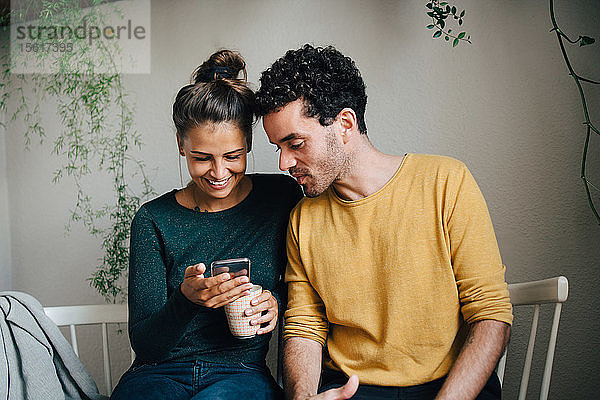 Lächelnde Freundin zeigt ihrem Freund ein Smartphone  während sie im Wohnzimmer Kaffee trinkt