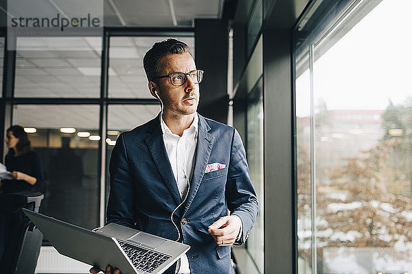 Nachdenklicher Unternehmer hält Laptop in der Hand  während er im Büro durchs Fenster schaut