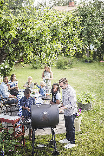 Schrägaufnahme eines Mannes  der mit einem Jungen einen Grill vorbereitet  während Familie und Freunde im Hinterhof am Tisch sitzen