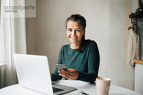 Porträt einer lächelnden mittelgroßen erwachsenen Frau mit Mobiltelefon und Laptop am Tisch