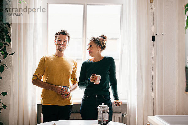 Glückliches Paar mit Kaffeetasse gegen Fenster zu Hause