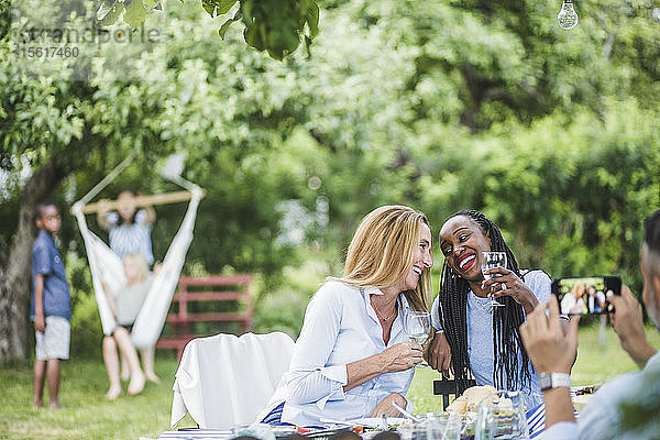 Mann mit Handy fotografiert lächelnde Frauen  die am Wochenende Wein im Garten feiern