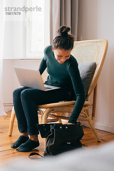 Lächelnde Frau in voller Länge mit Laptop  die in die Handtasche schaut  während sie zu Hause auf einem Stuhl sitzt