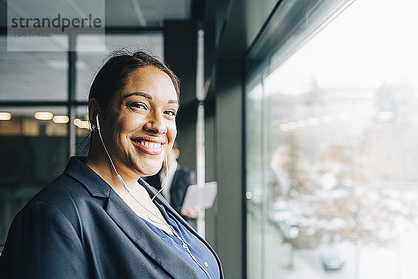 Porträt einer lächelnden Geschäftsfrau mit Kopfhörern  die im Büro am Fenster steht