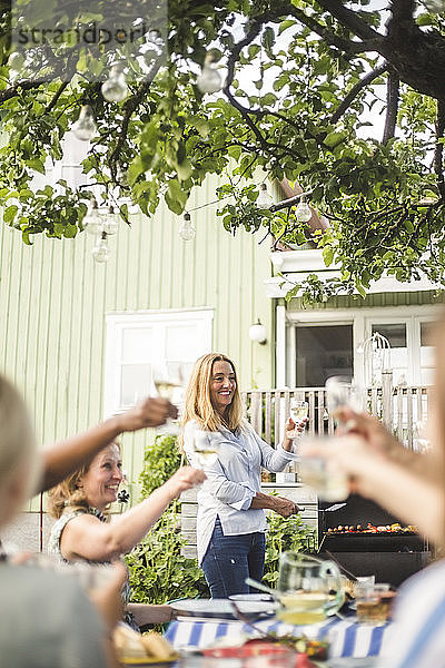 Reife Frau bereitet Essen auf dem Grill zu  während sie auf einer Gartenparty mit Freunden auf Weingläser anstößt