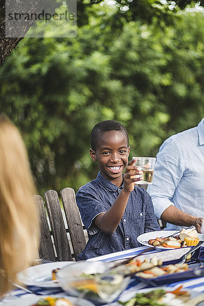 Lächelnder Junge hält Trinkglas in der Hand  während er während der Party mit seiner Familie im Garten sitzt