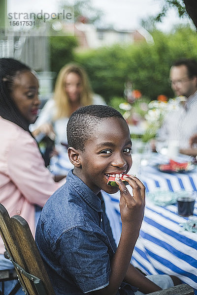 Lächelnder Junge isst Wassermelone  während er mit der Familie am Tisch im Garten sitzt