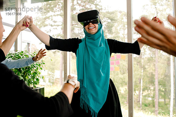 Fröhliche Geschäftsfrau hält die Hand ihres Kollegen  während sie den Virtual-Reality-Simulator während des Kick-off-Meetings bei der Versammlung benutzt