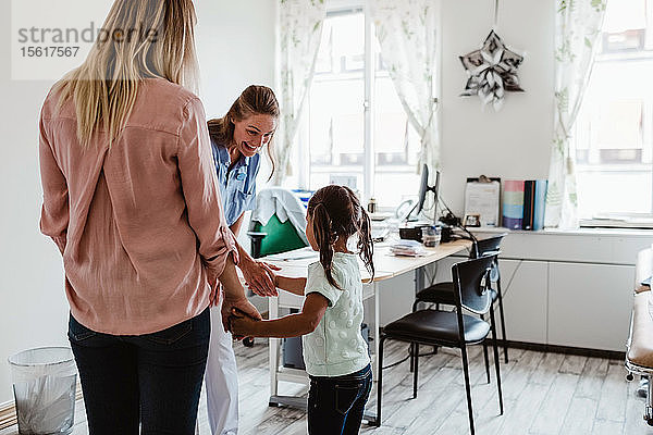 Lächelnder Kinderarzt schüttelt einem Mädchen die Hand  während die Mutter in der Arztpraxis steht