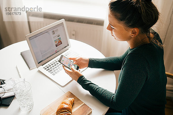 Frau benutzt Smartphone  während sie zu Hause mit ihrem Laptop auf einem Lebensmittelrezept surft