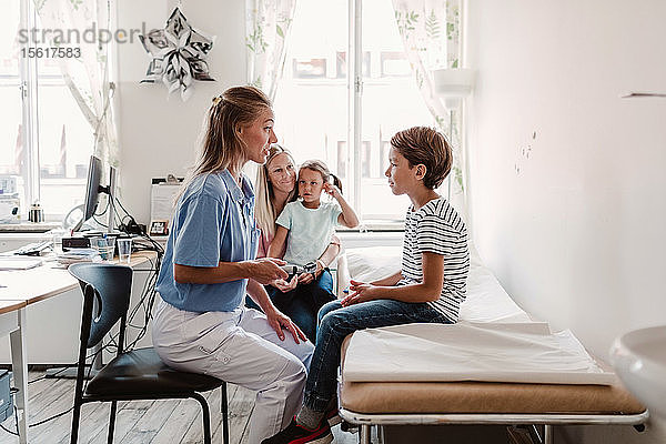 Weibliche Kinderärztin mit Otoskop  die den Jungen untersucht  während die Familie im Hintergrund in der Klinik sitzt
