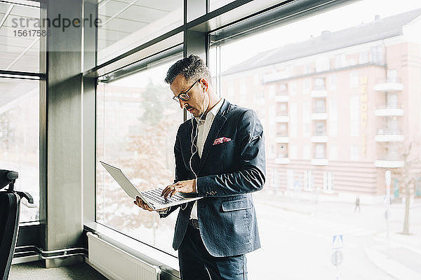 Reifer Geschäftsmann benutzt Laptop  während er im Büro vor dem Fenster steht