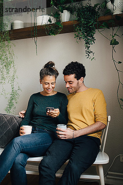 Lächelnder Freund und Freundin  die beim Kaffee im Wohnzimmer auf das Handy schauen