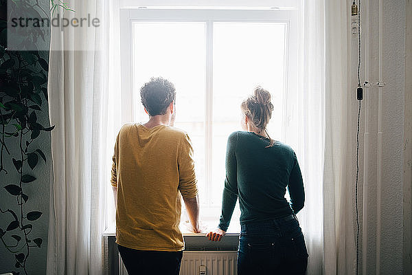 Rückansicht eines zu Hause stehenden Paares  das durch ein Fenster schaut