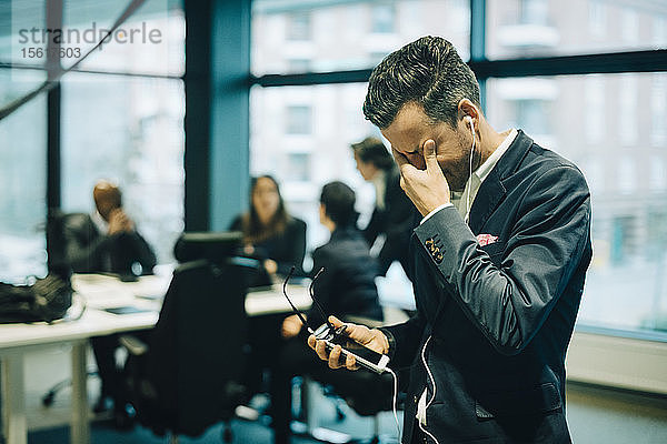 Gestresster Geschäftsmann benutzt Telefon über Kopfhörer  während er sich im Büro die Augen reibt