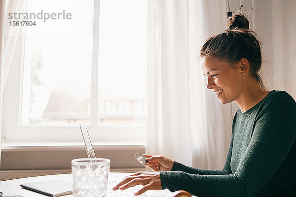 Seitenansicht einer lächelnden Frau mit Kreditkarte am Laptop im Wohnzimmer