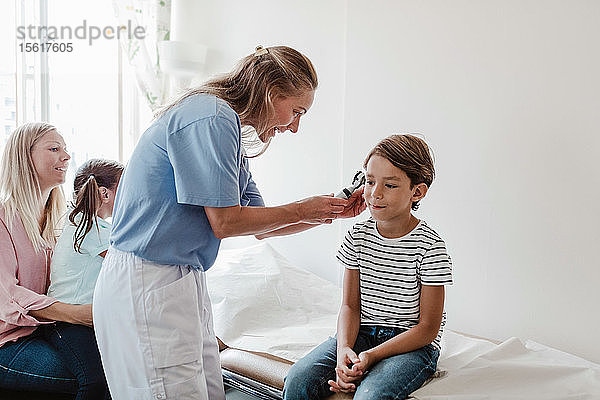 Weibliche Ärztin untersucht das Ohr des Jungen mit dem Otoskop  während die Familie im Behandlungszimmer sitzt