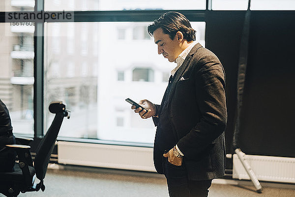 Seitenansicht eines Geschäftsmannes  der ein Mobiltelefon benutzt  während er am Arbeitsplatz steht