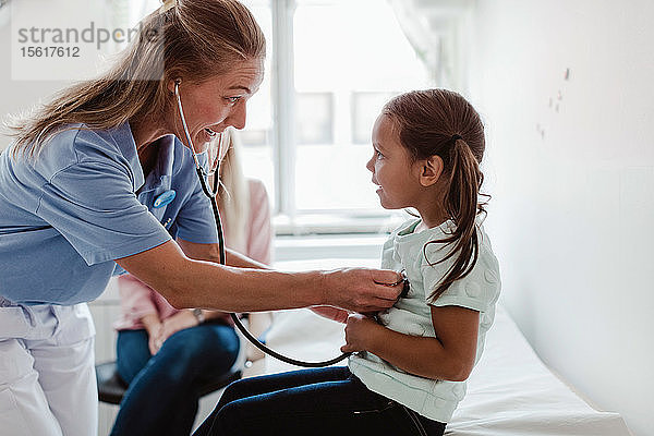 Weibliche Kinderärztin untersucht den Herzschlag des Mädchens durch ein Stethoskop  während die Mutter im Hintergrund in der Klinik sitzt