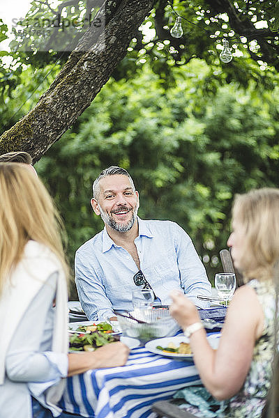 Glücklicher Mann genießt mit Freunden am Esstisch bei einer Gartenparty